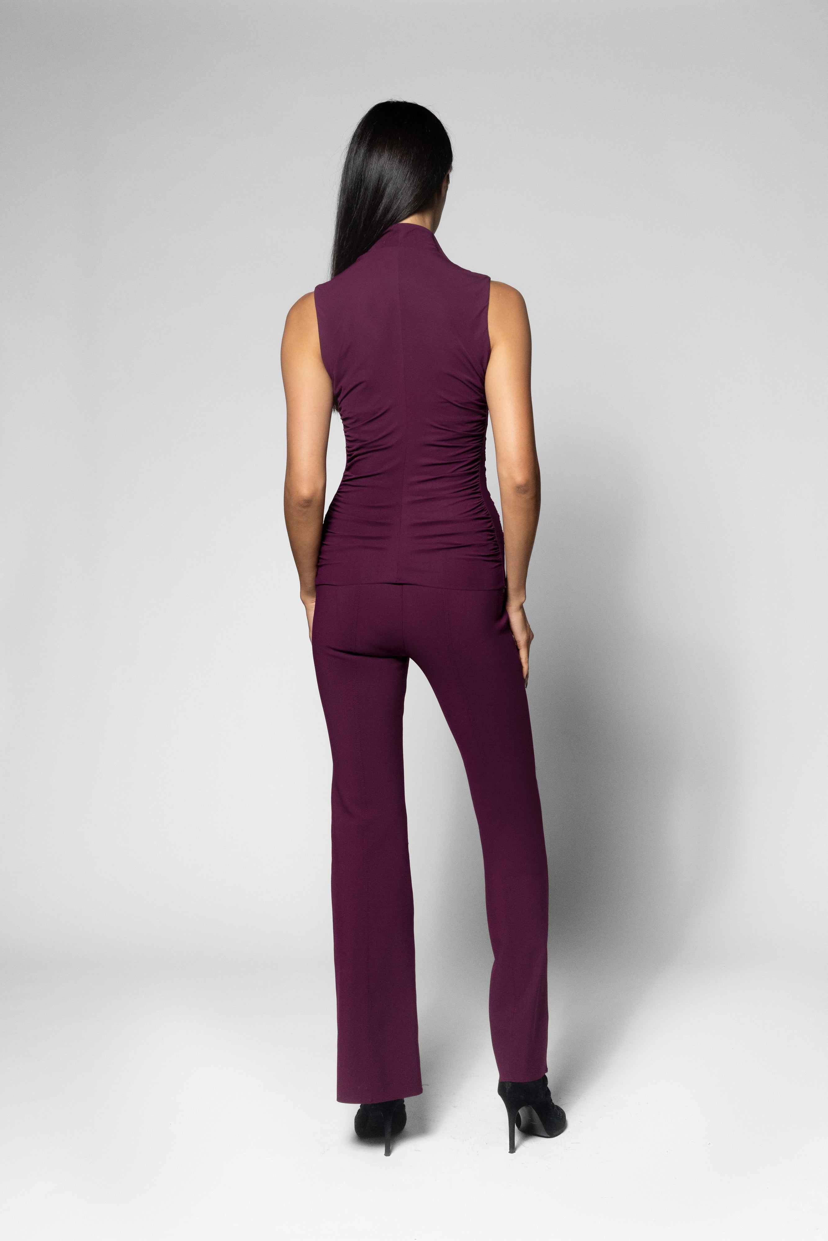 A woman viewed from the back wearing a sleeveless high-neck shirt in deep plum purple with matching pants, paired with black heels, against a light gray studio background.