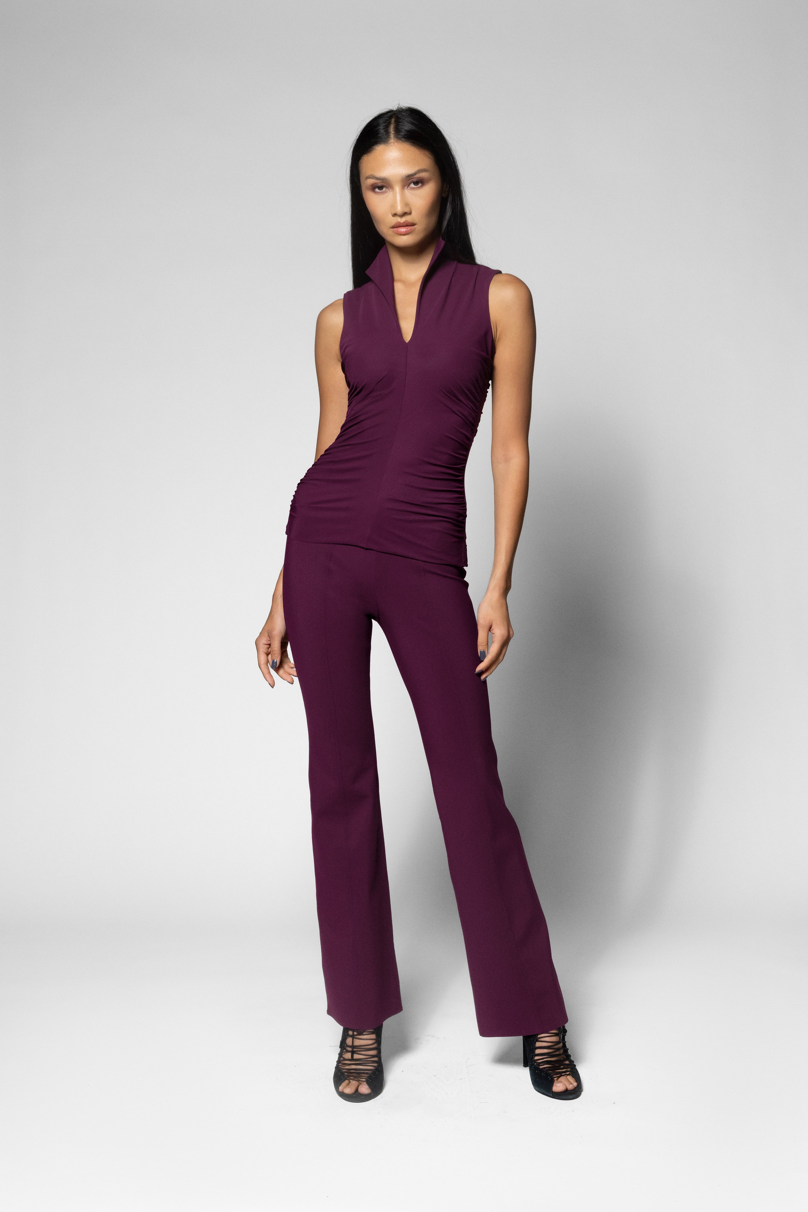 A woman modeling a sleeveless, deep purple shirt with a wrap-style V-neck and straight-leg matching pants, paired with black strappy high-heeled sandals, against a light gray studio background.