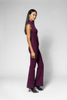 A woman modeling a sleeveless, high-neck, dark purple shirt with a ruched bodice and wide-leg pants, standing against a light gray studio background.