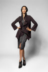 A model poses in a dark plum-colored wrap jacket over a textured gray and black patterned dress, paired with black pointed-toe heels, against a light gray studio background.