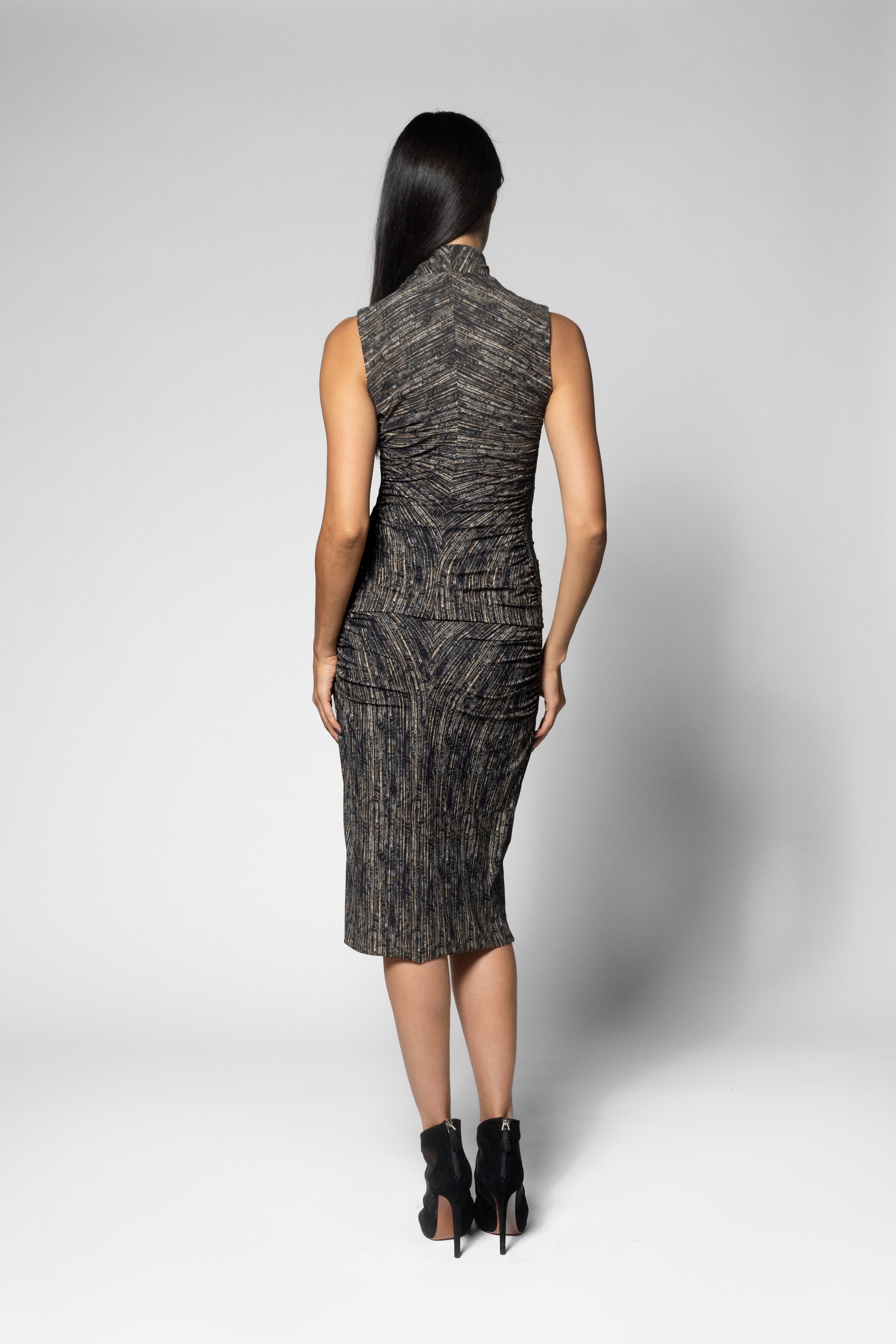 A woman viewed from the back wearing a sleeveless, high-neck top with matching knee-length skirt with a textured black and gray pattern, paired with black ankle boots, against a light gray studio background.
