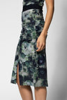A woman wearing a ruched midi skirt with a green, blue, and gray watercolor floral print, featuring a side slit and a black waistband.