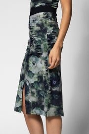 A woman wearing a ruched midi skirt with a green, blue, and gray watercolor floral print, featuring a side slit and a black waistband.