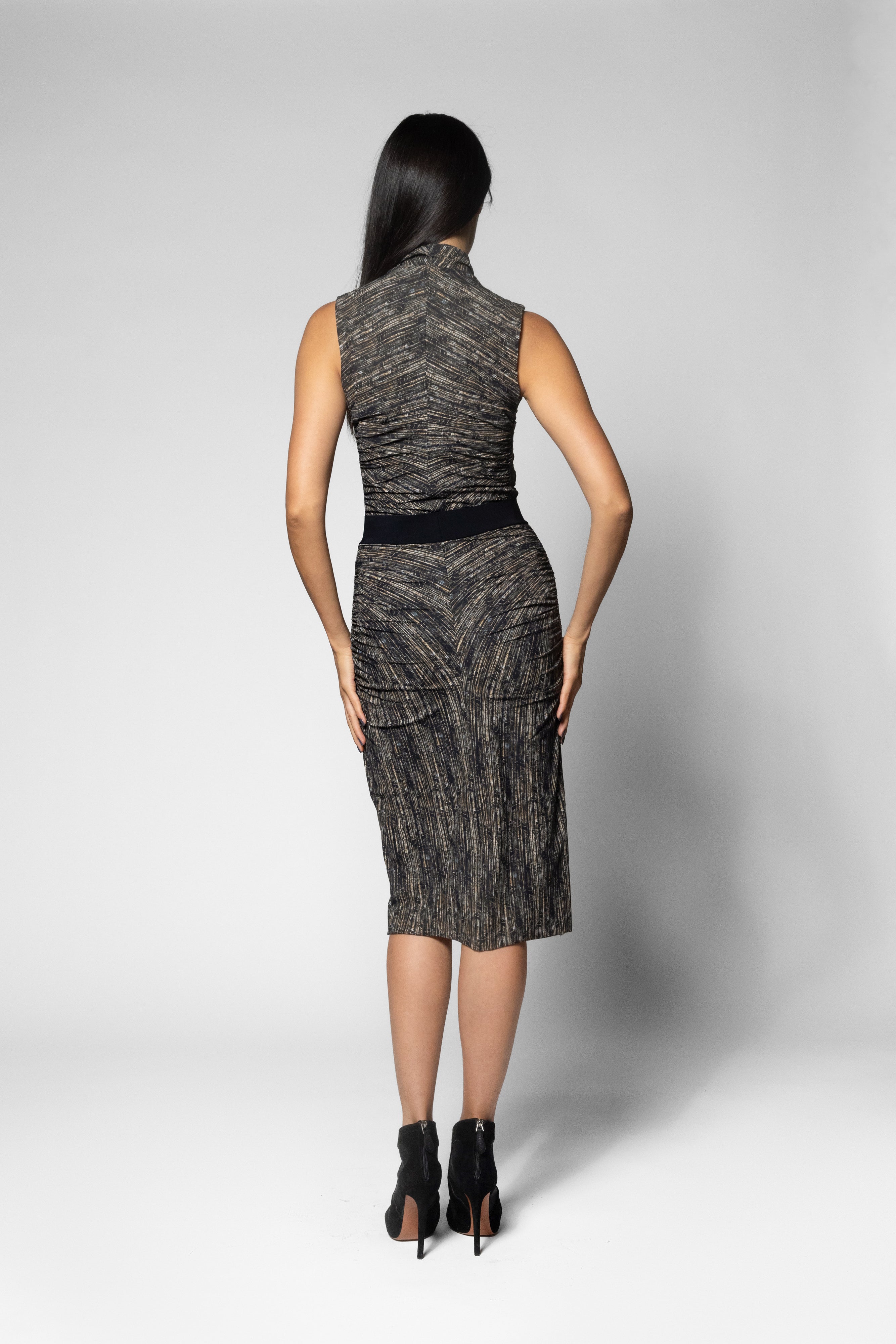 A woman viewed from the back wearing a sleeveless high-neck shirt and matching printed skirt with a textured black and gray pattern, a black waistband, and black ankle boots, set against a light gray studio background.