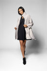A model wearing a light beige oversized wrap coat over a black dress and black ankle boots, posing against a light gray studio background.