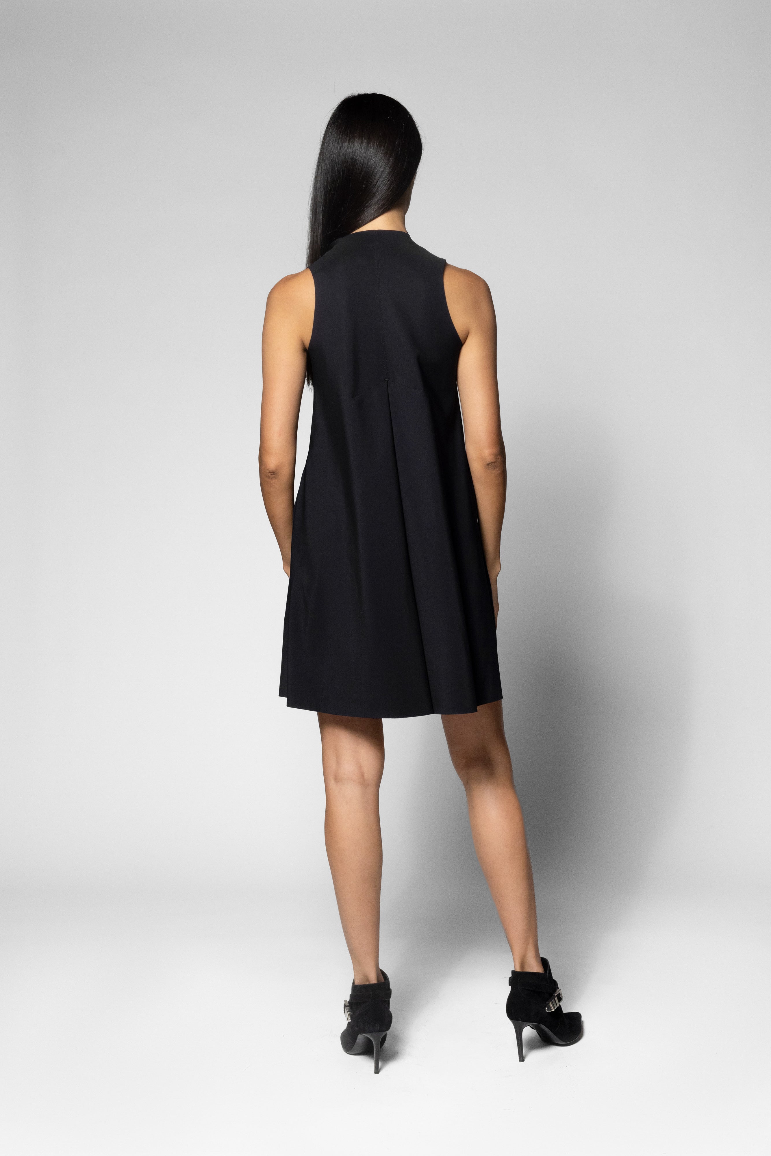 A woman viewed from behind wearing a sleeveless black mini dress and black high heels against a light gray studio background.
