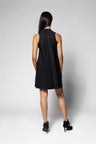 A woman viewed from behind wearing a sleeveless black mini dress and black high heels against a light gray studio background.