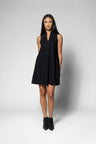 A woman modeling a sleeveless black mini dress with a V-neckline, standing against a light gray studio background.