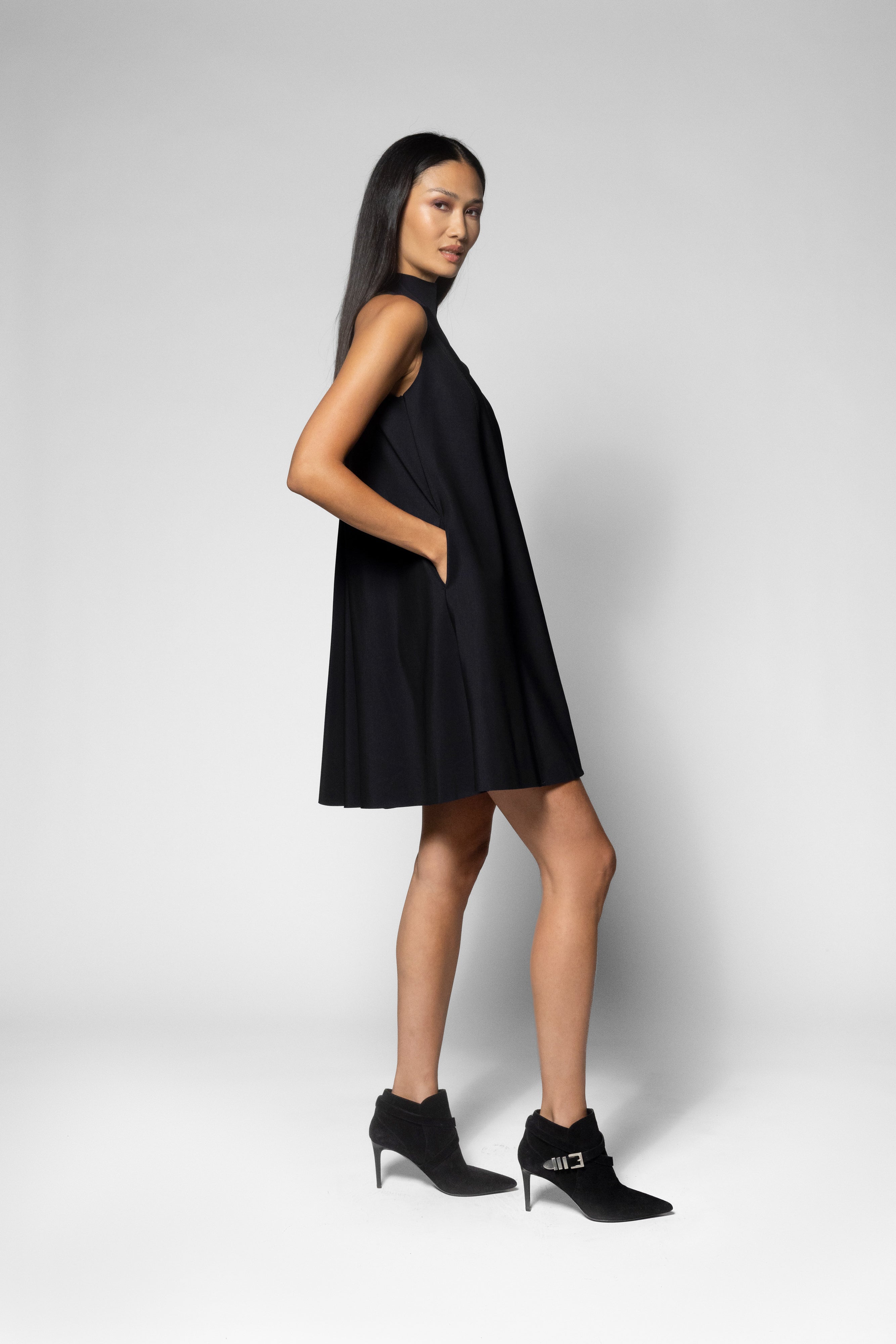 A woman modeling a sleeveless black mini dress with a high neckline and side pockets, paired with black ankle boots, against a light gray studio background.