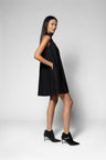 A woman modeling a sleeveless black mini dress with a high neckline and side pockets, paired with black ankle boots, against a light gray studio background.