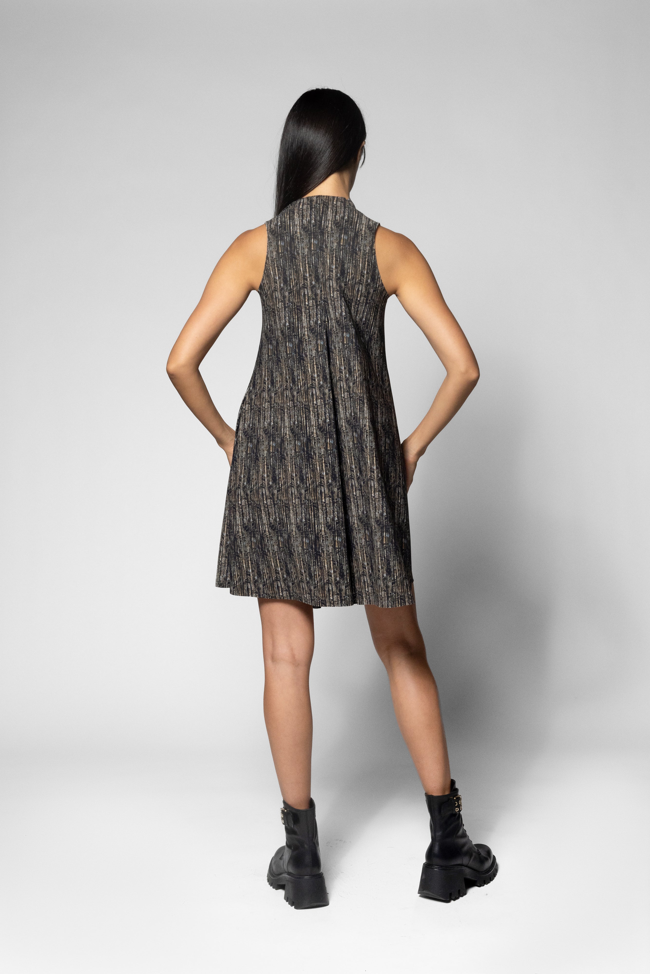 A woman is shown from the back wearing a sleeveless, knee-length pleated dress with a textured black and brown pattern, paired with black combat boots, against a light gray studio background.