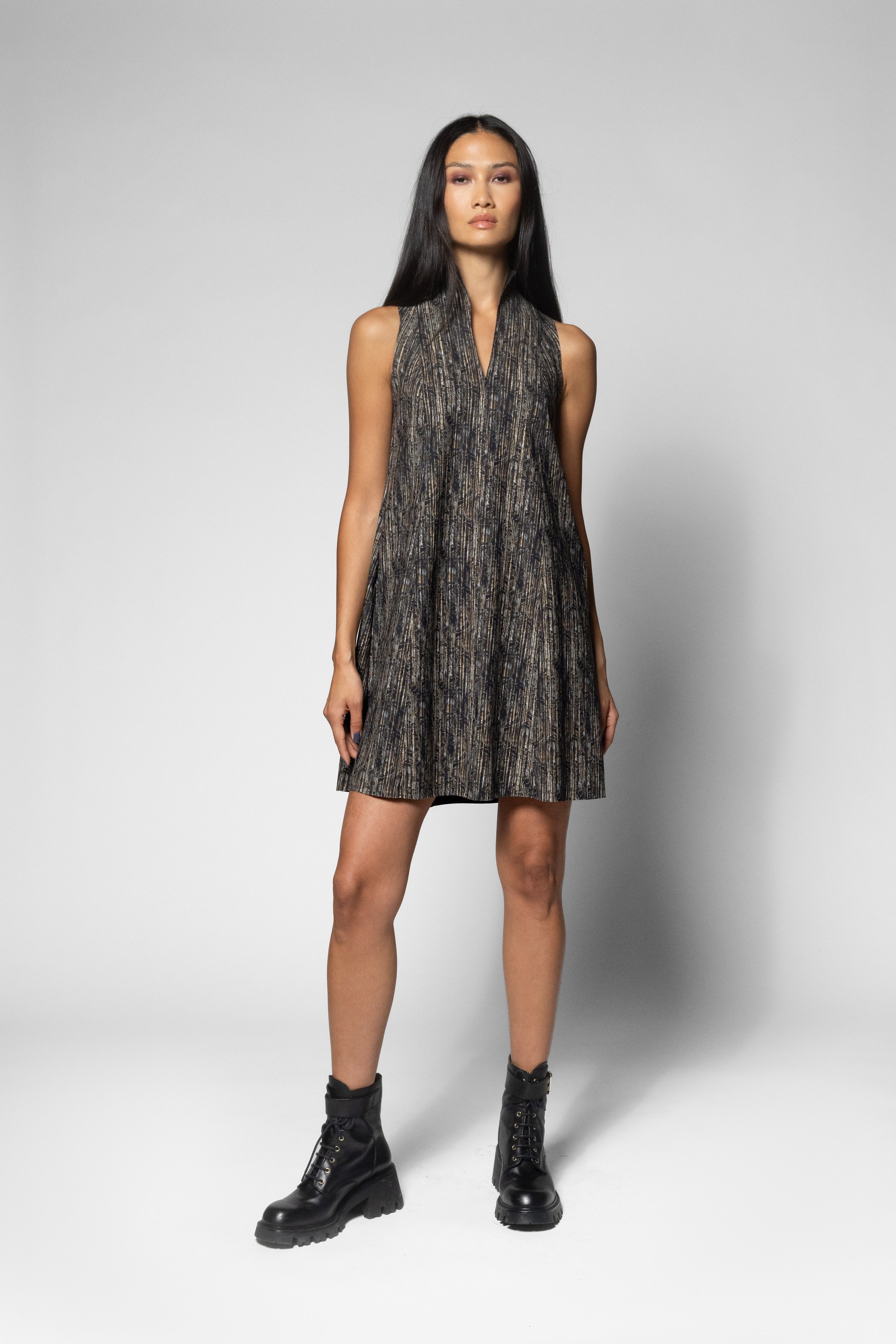 A model stands against a light gray studio background wearing a sleeveless V-neck mini dress with a dark abstract pattern and black combat boots.