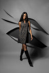 A woman modeling a sleeveless black and gray patterned mini dress with black knee-high boots, standing against a dark gray studio backdrop with abstract black ribbon-like shapes.