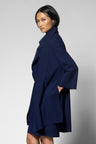 A woman modeling a navy blue wrap-style coat with wide sleeves and a high collar, photographed against a plain light gray studio background.