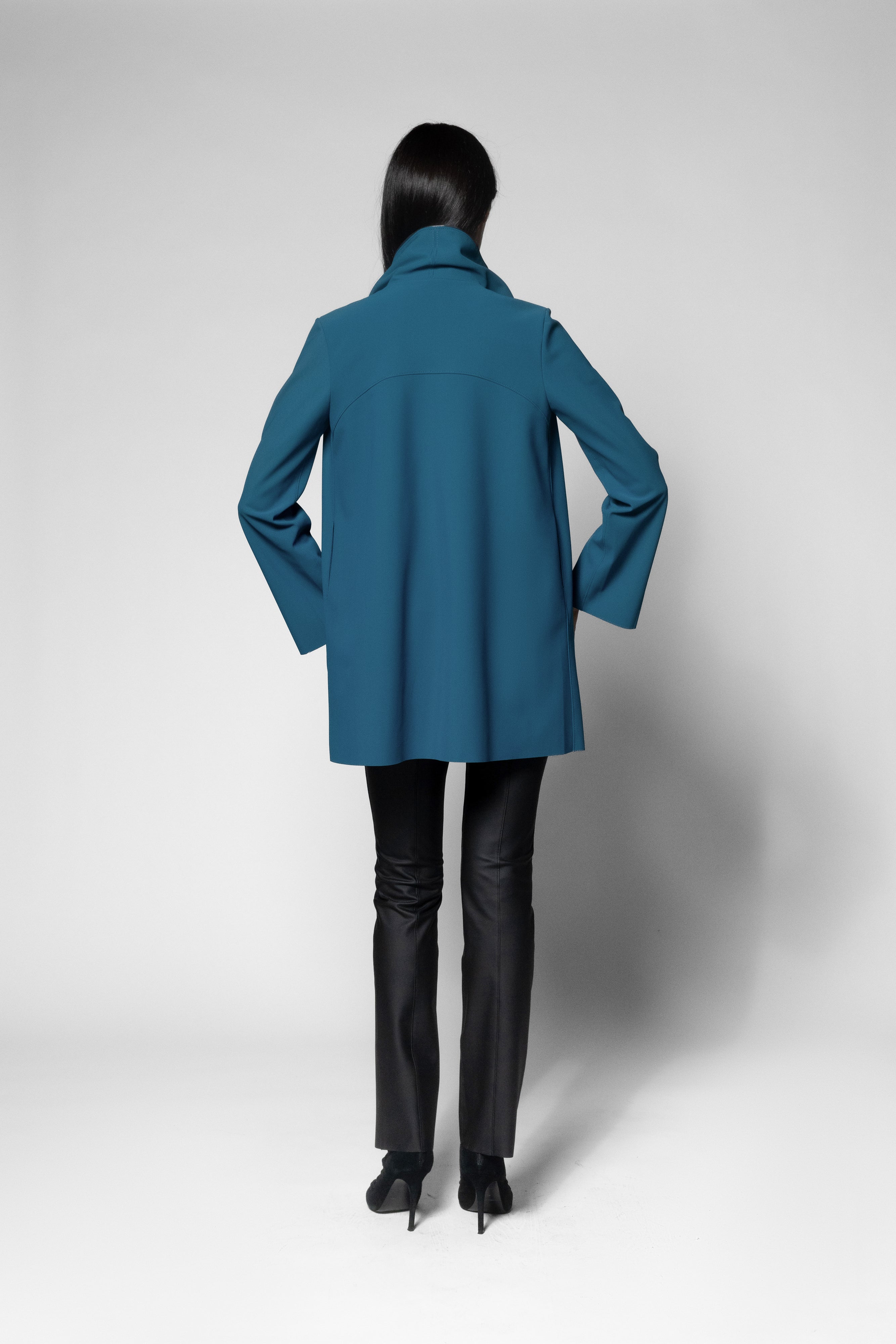 A model shown from the back wearing a teal blue coat and black pants against a light gray studio background.