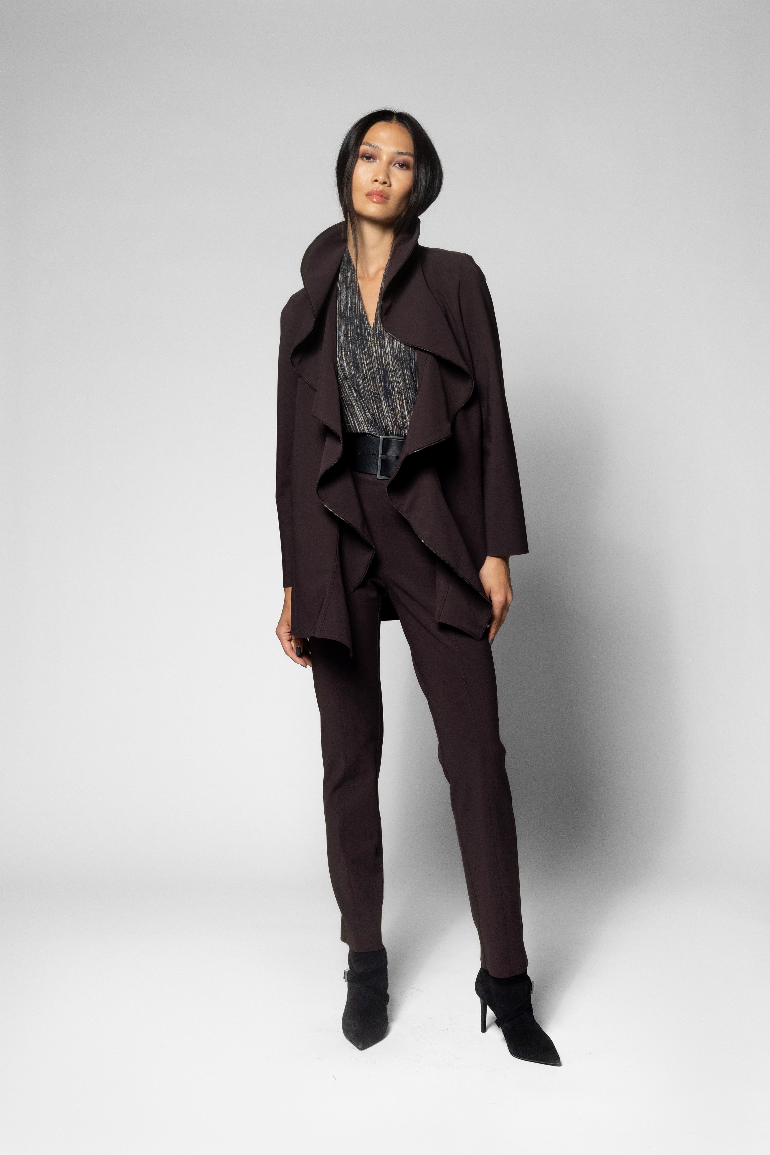 A model stands in a studio wearing a dark plum-brown tailored set consisting of a draped jacket, matching trousers, and a patterned top, paired with black pointed-toe heels.