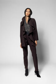 A model stands in a studio wearing a dark plum-brown tailored set consisting of a draped jacket, matching trousers, and a patterned top, paired with black pointed-toe heels.