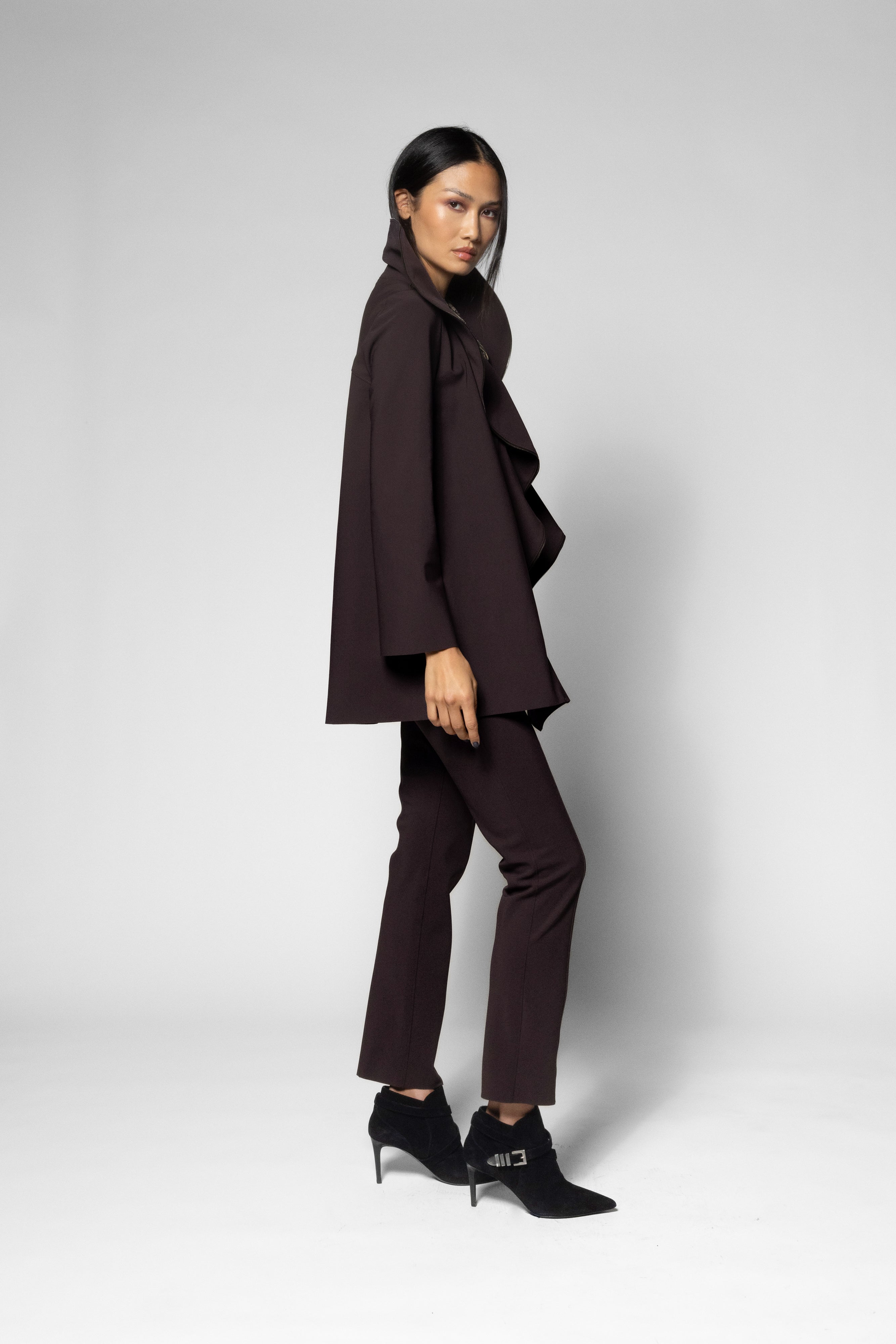 A model is shown in profile wearing a dark brown ruffled cape-style jacket, matching dark brown trousers, and black ankle boots against a light gray studio background.