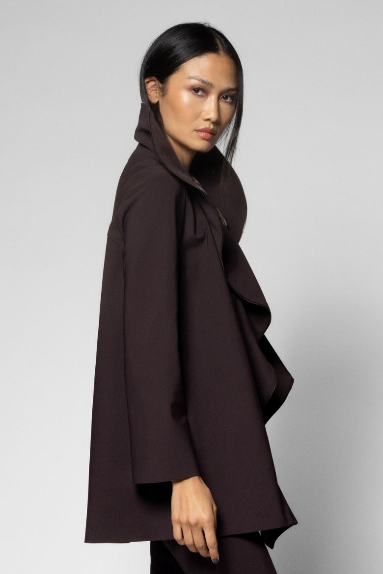 A model wearing a dark brown, draped, ruffled coat with a high collar, photographed against a neutral light gray studio background.