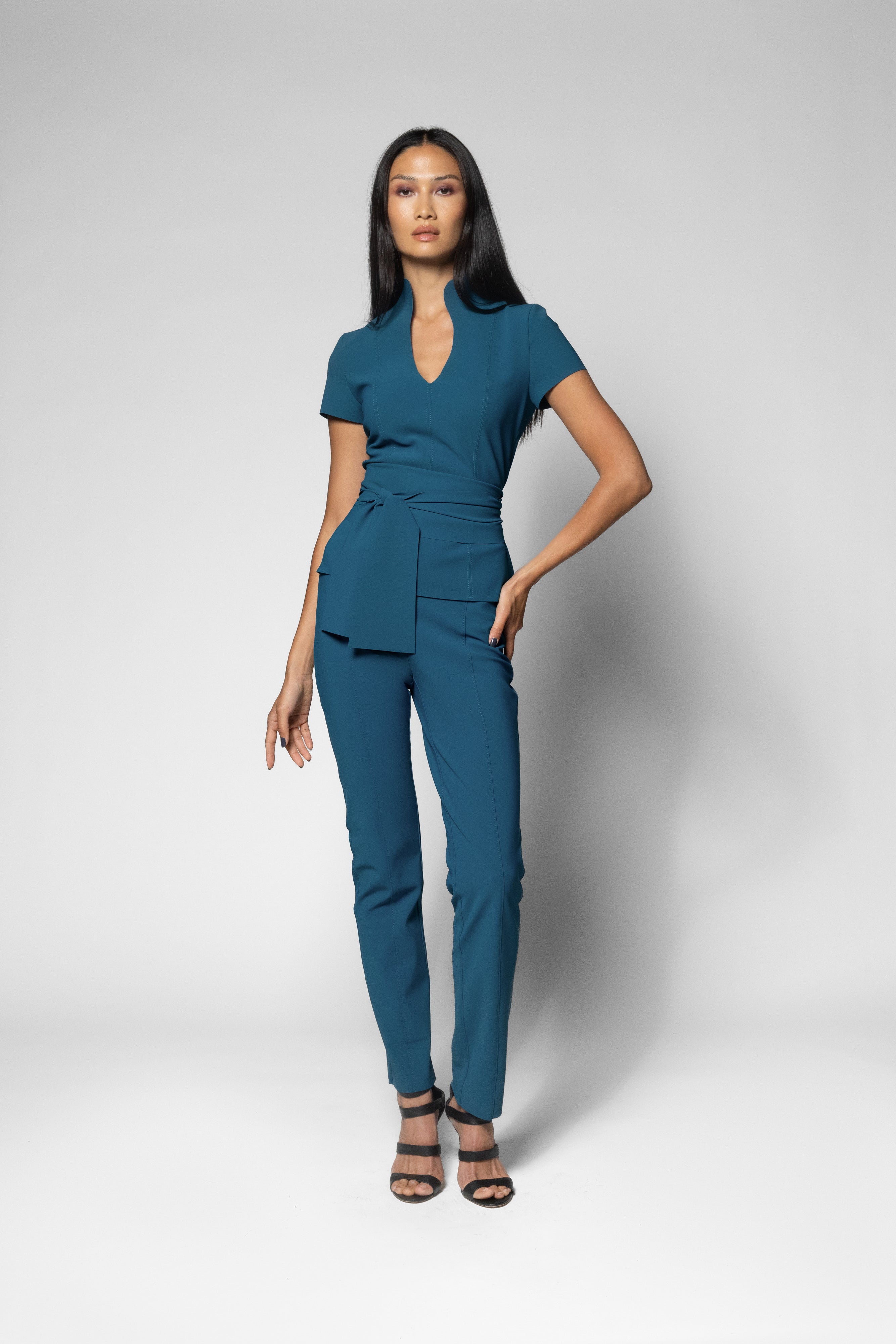 A woman modeling a teal blue wrap-style short-sleeve top and matching slim-fit trousers, styled with black strappy heels against a light gray studio background.