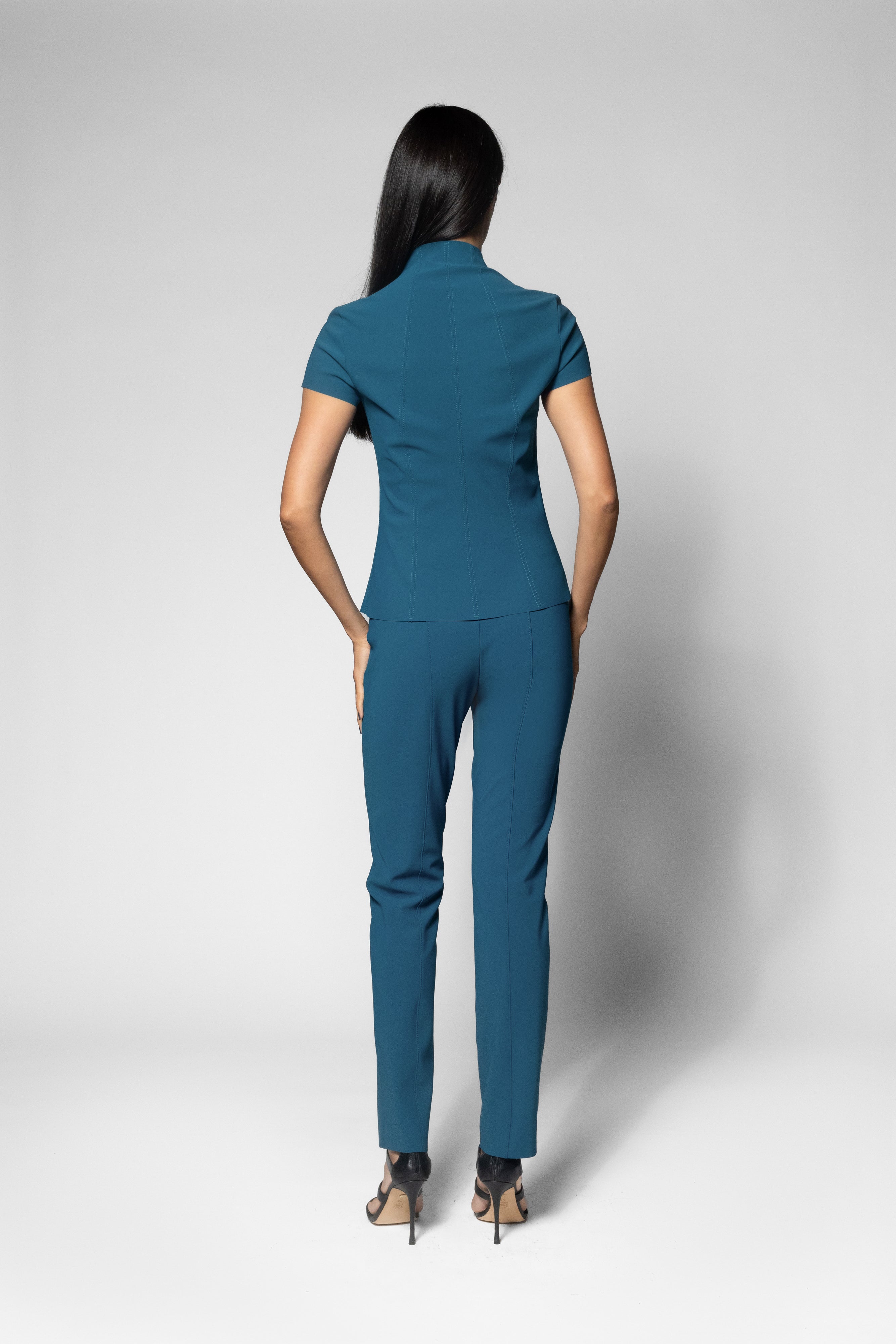 A woman viewed from the back wearing a teal short-sleeve collared top and matching slim trousers, standing against a light gray studio background.