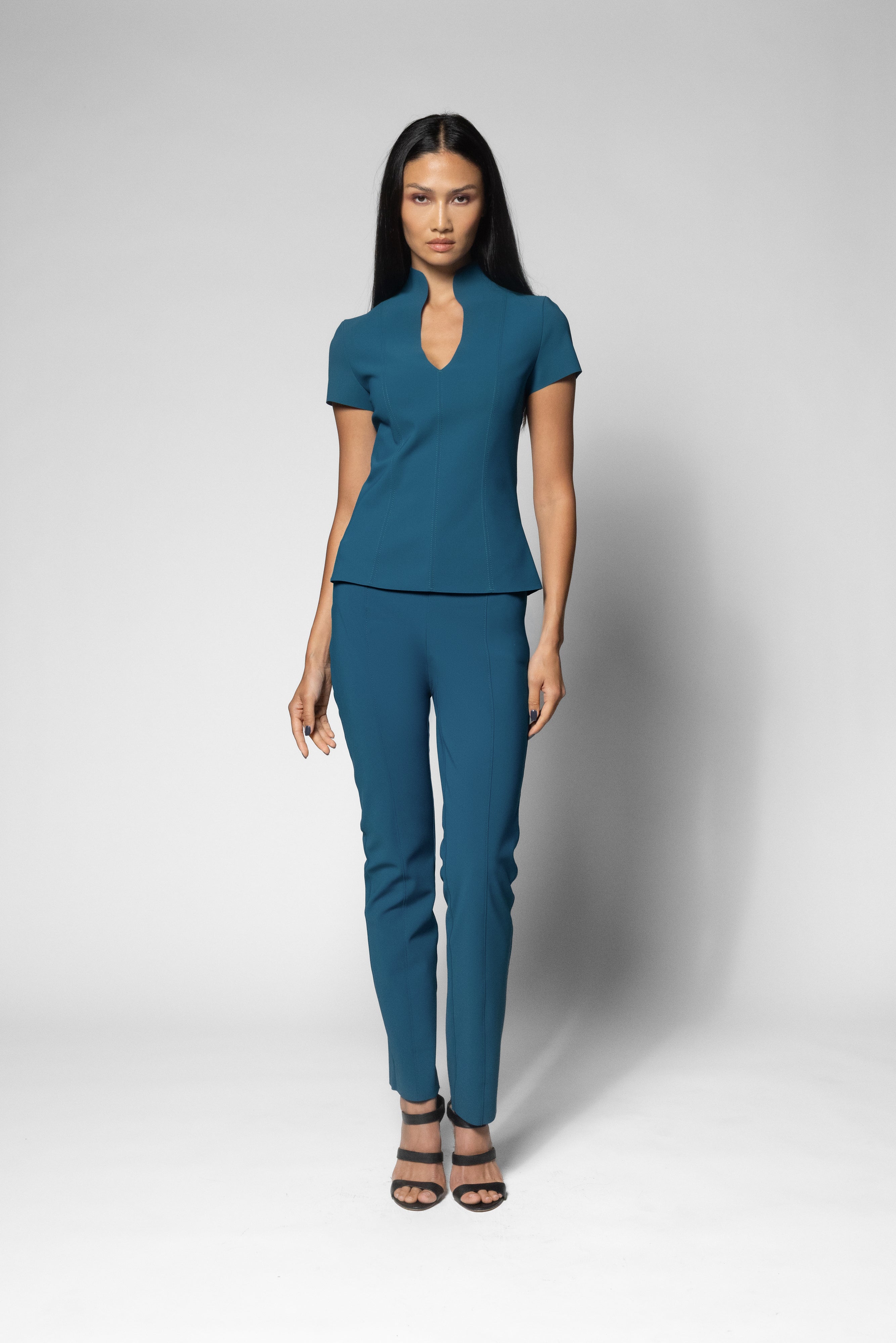 A woman modeling a matching teal blue two-piece set consisting of a short-sleeve peplum top with a keyhole neckline and slim-fit trousers, paired with black strappy heels, against a light gray studio background.