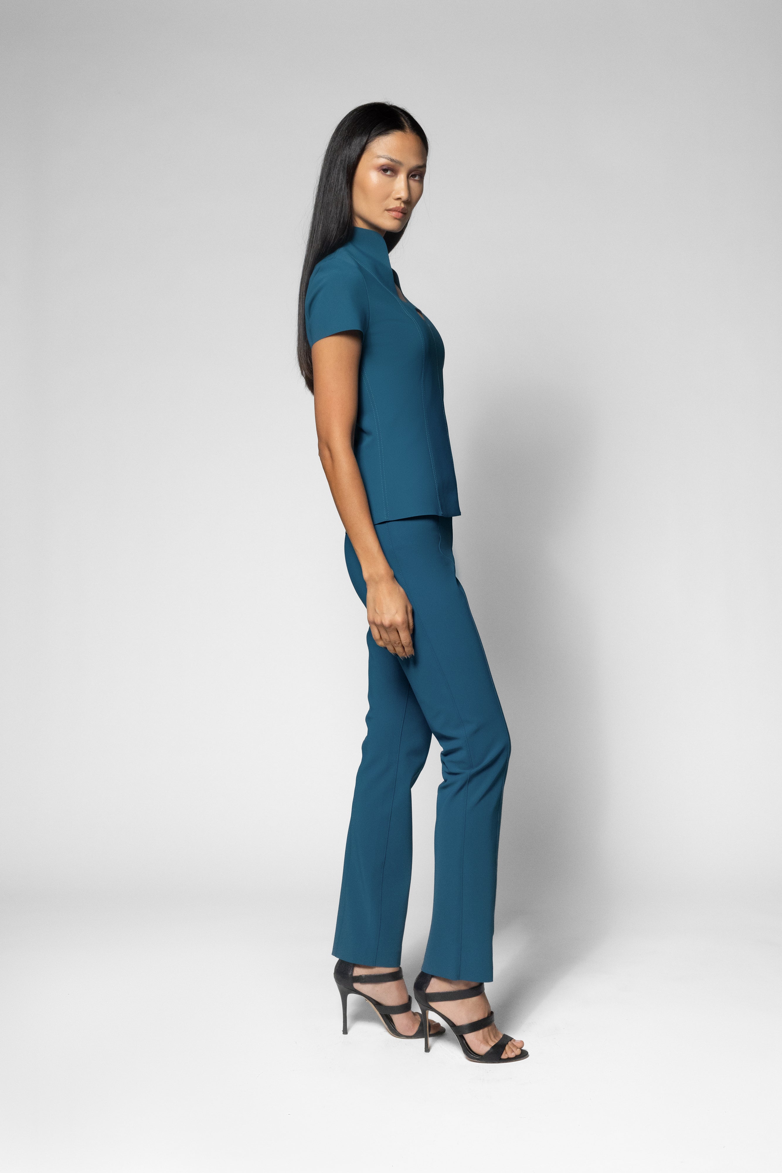 A woman modeling a teal blue two-piece set consisting of a short-sleeve mock-neck top and matching wide-leg trousers, paired with black strappy high-heeled sandals against a light gray studio background.