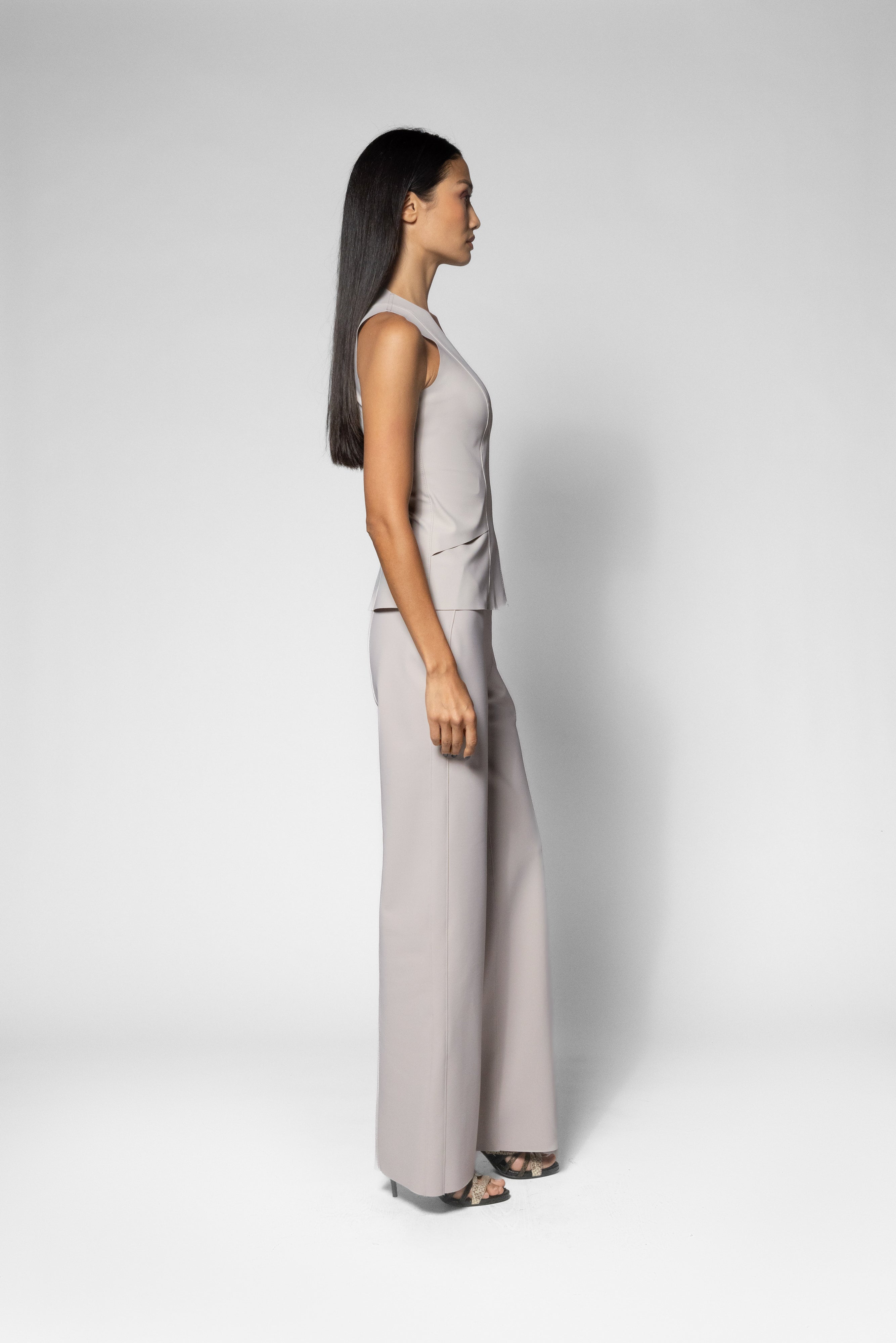 A woman in a side profile wearing a light beige sleeveless top and matching wide-leg pants, standing against a neutral light gray studio background.
