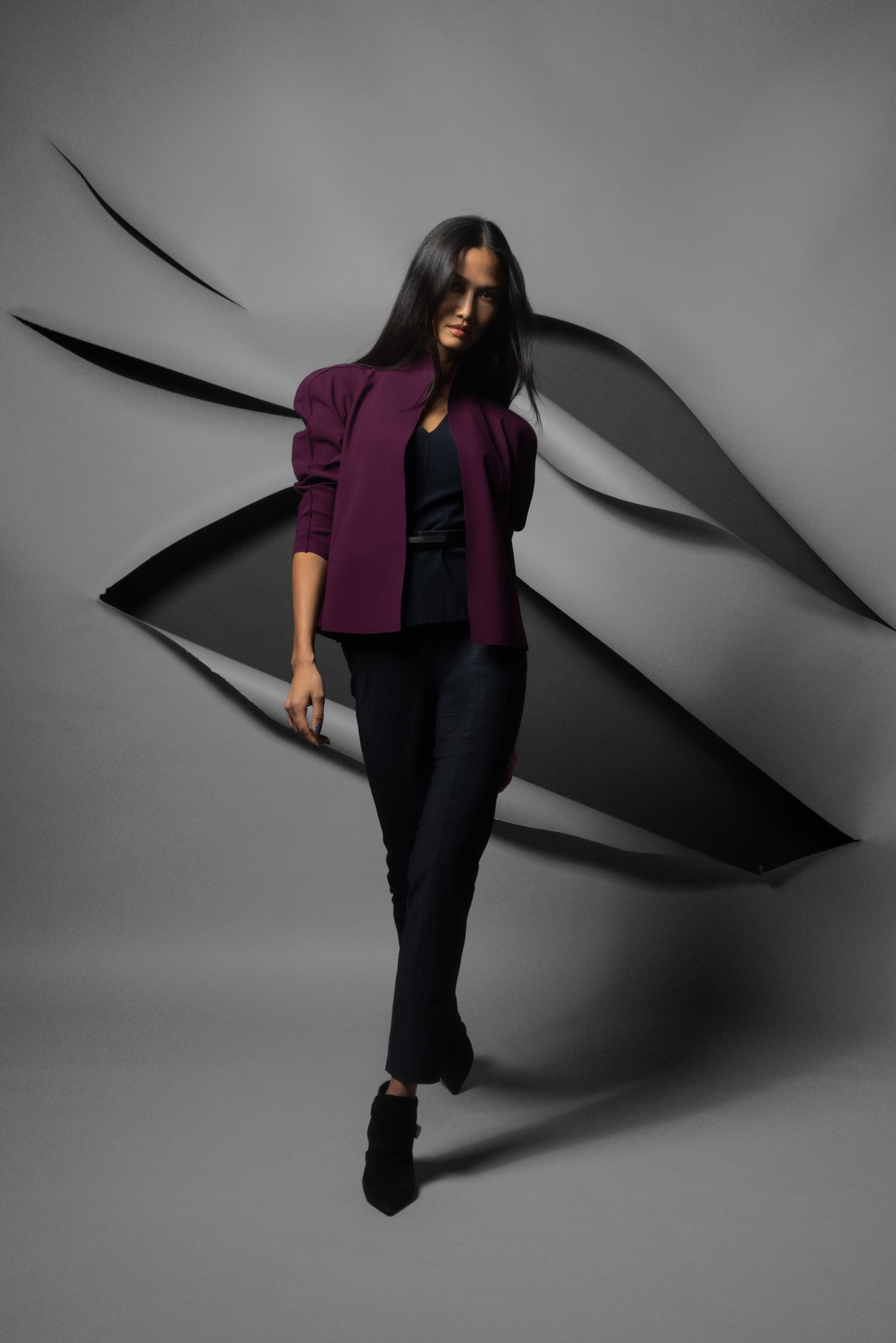 A woman modeling a dark plum purple blazer, black top, and black trousers against a gray studio backdrop with abstract black layered shapes behind her.