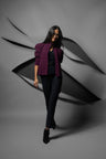 A woman modeling a dark plum purple blazer, black top, and black trousers against a gray studio backdrop with abstract black layered shapes behind her.