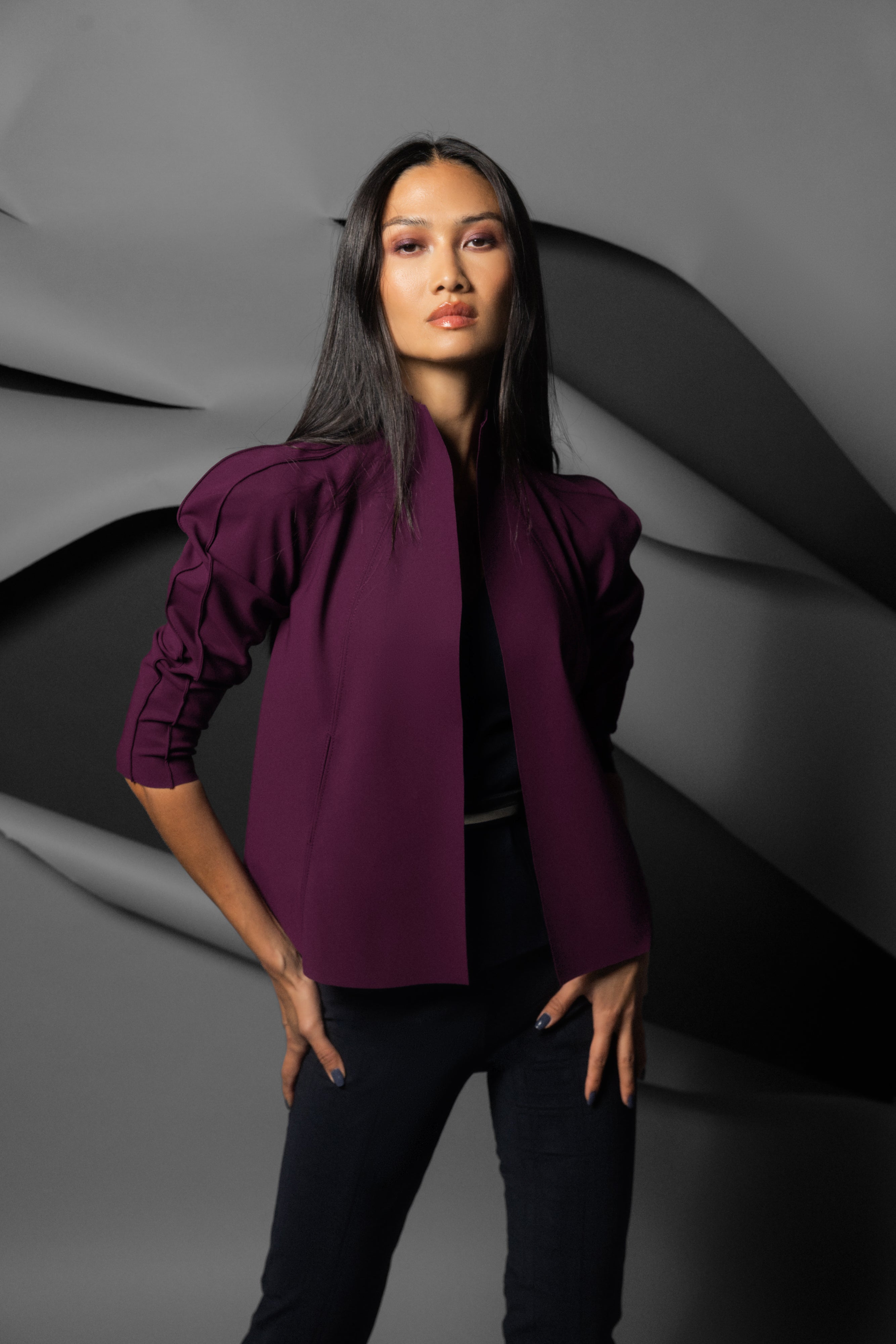 A woman modeling a structured, deep purple cropped jacket with puffed shoulders, worn over a black top and black pants, set against a dark gray abstract background.