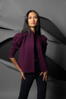 A woman modeling a structured, deep purple cropped jacket with puffed shoulders, worn over a black top and black pants, set against a dark gray abstract background.