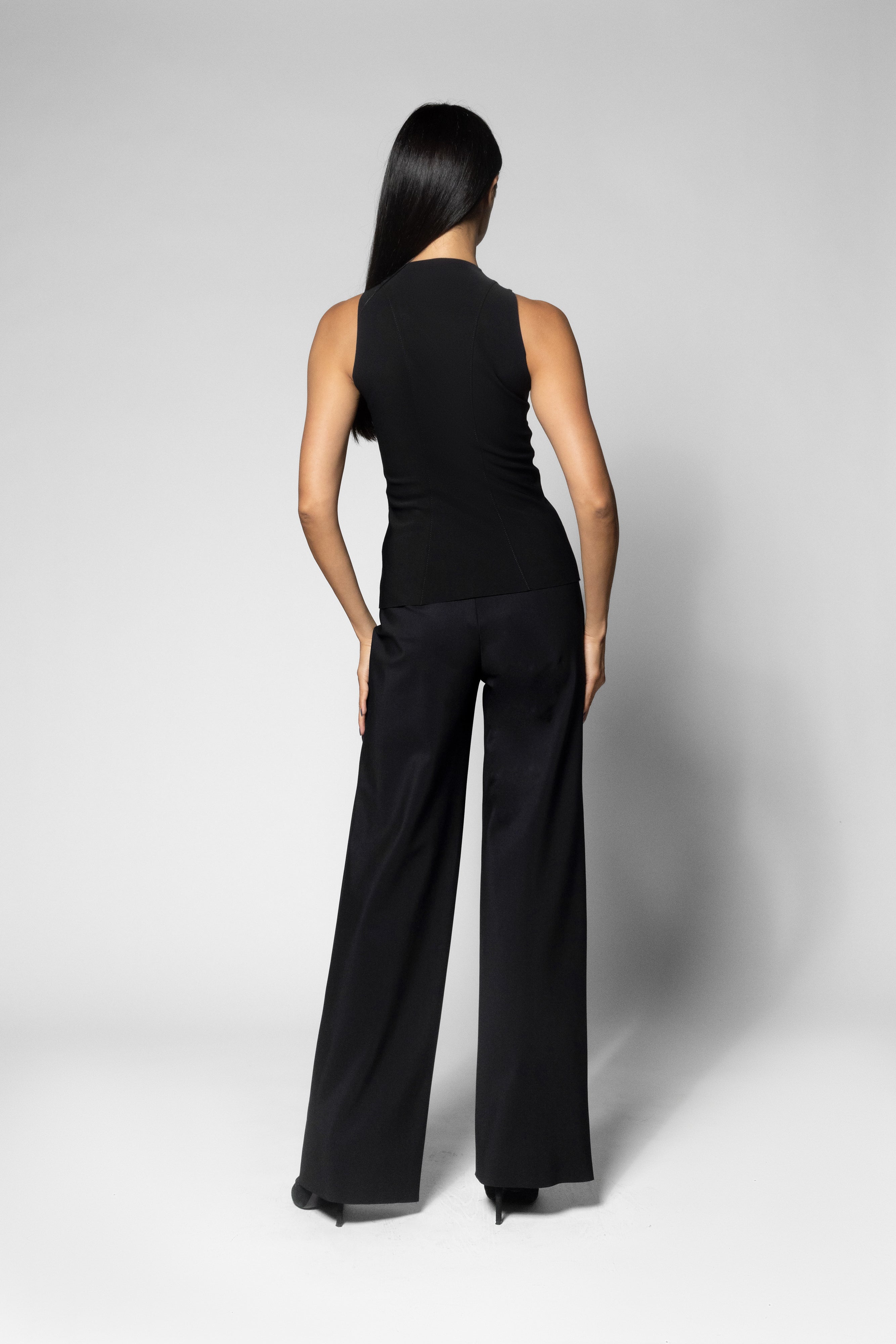 A woman is shown from the back wearing a black sleeveless top and black wide-leg trousers against a light gray studio background.