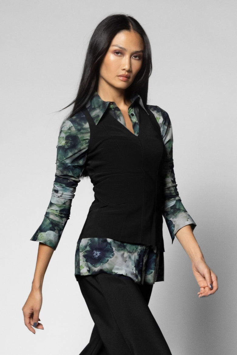 A woman modeling a black vest and long-sleeve blouse with green and blue floral watercolor patterned sleeves, collar, and hem, paired with black pants against a light gray studio background.