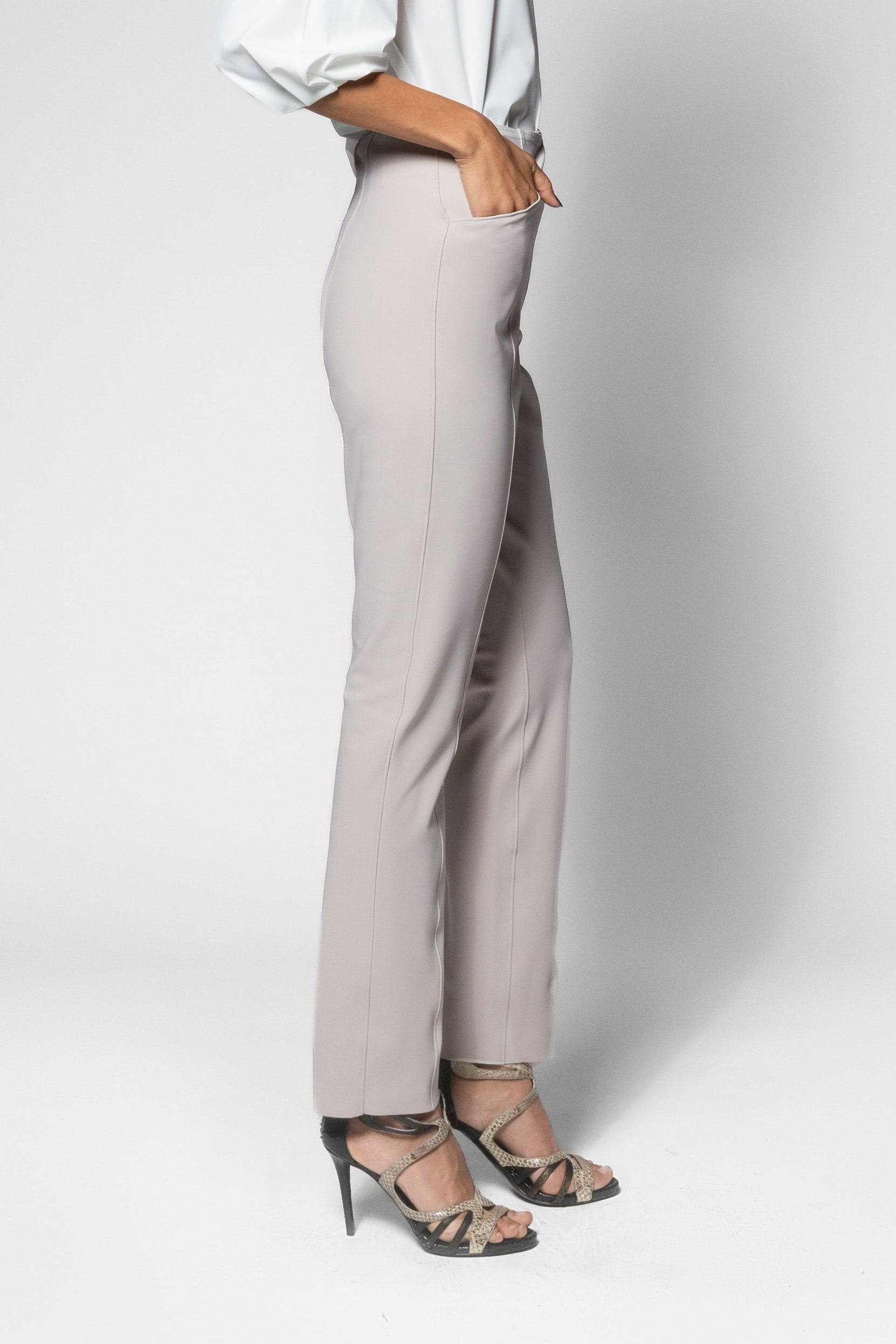 A woman stands in profile wearing a white puff-sleeve blouse and light beige straight-leg trousers with strappy high-heeled sandals, photographed against a light gray studio background.