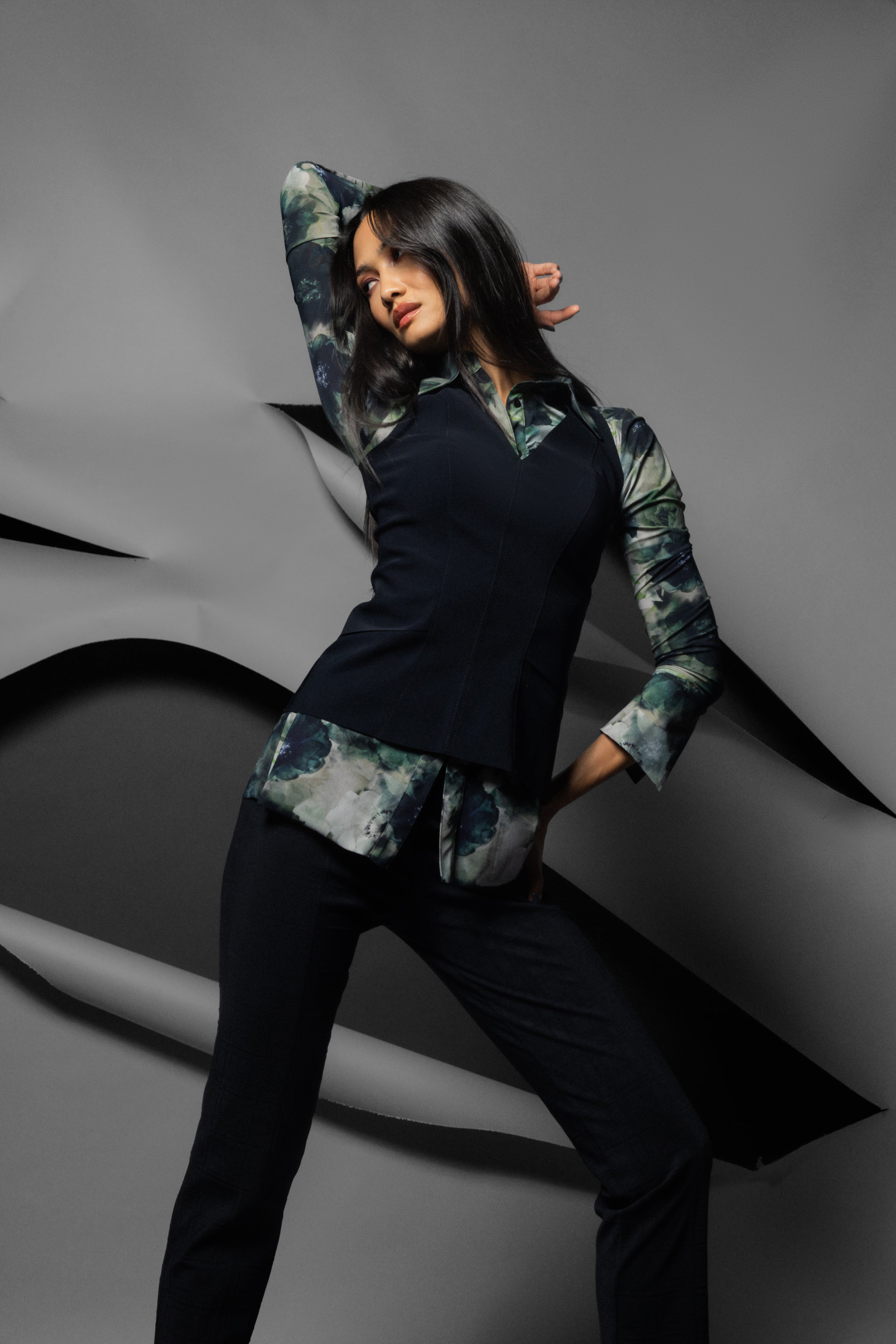 A woman poses in a studio wearing a long-sleeve collared shirt with a black body and green and gray abstract patterned sleeves, paired with dark trousers, against a gray background with abstract curved shapes.