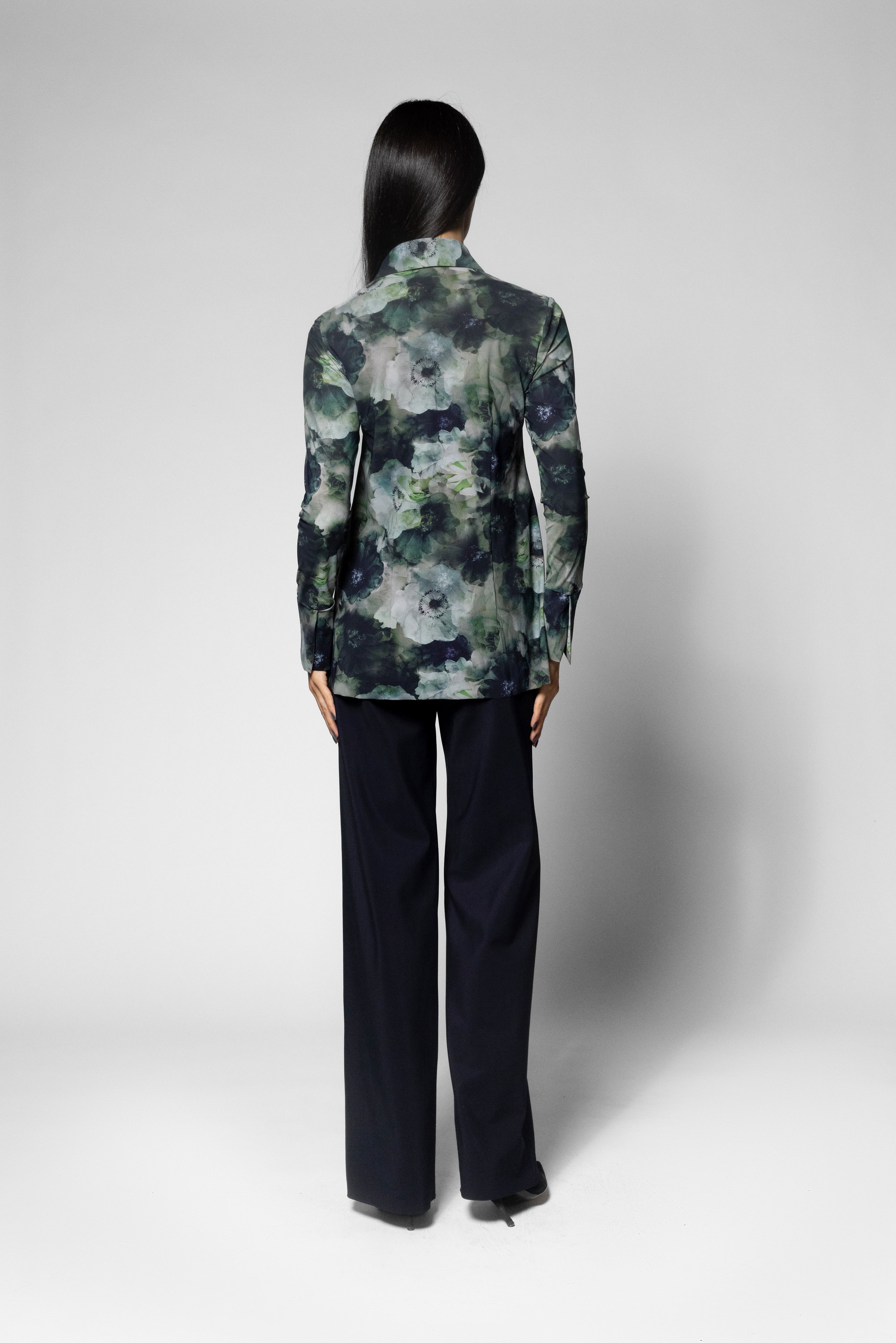 A model shown from the back wearing a long-sleeve collared blazer with a green, gray, and black abstract watercolor pattern, paired with dark navy wide-leg trousers, against a light gray studio background.