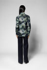 A model shown from the back wearing a long-sleeve collared blazer with a green, gray, and black abstract watercolor pattern, paired with dark navy wide-leg trousers, against a light gray studio background.