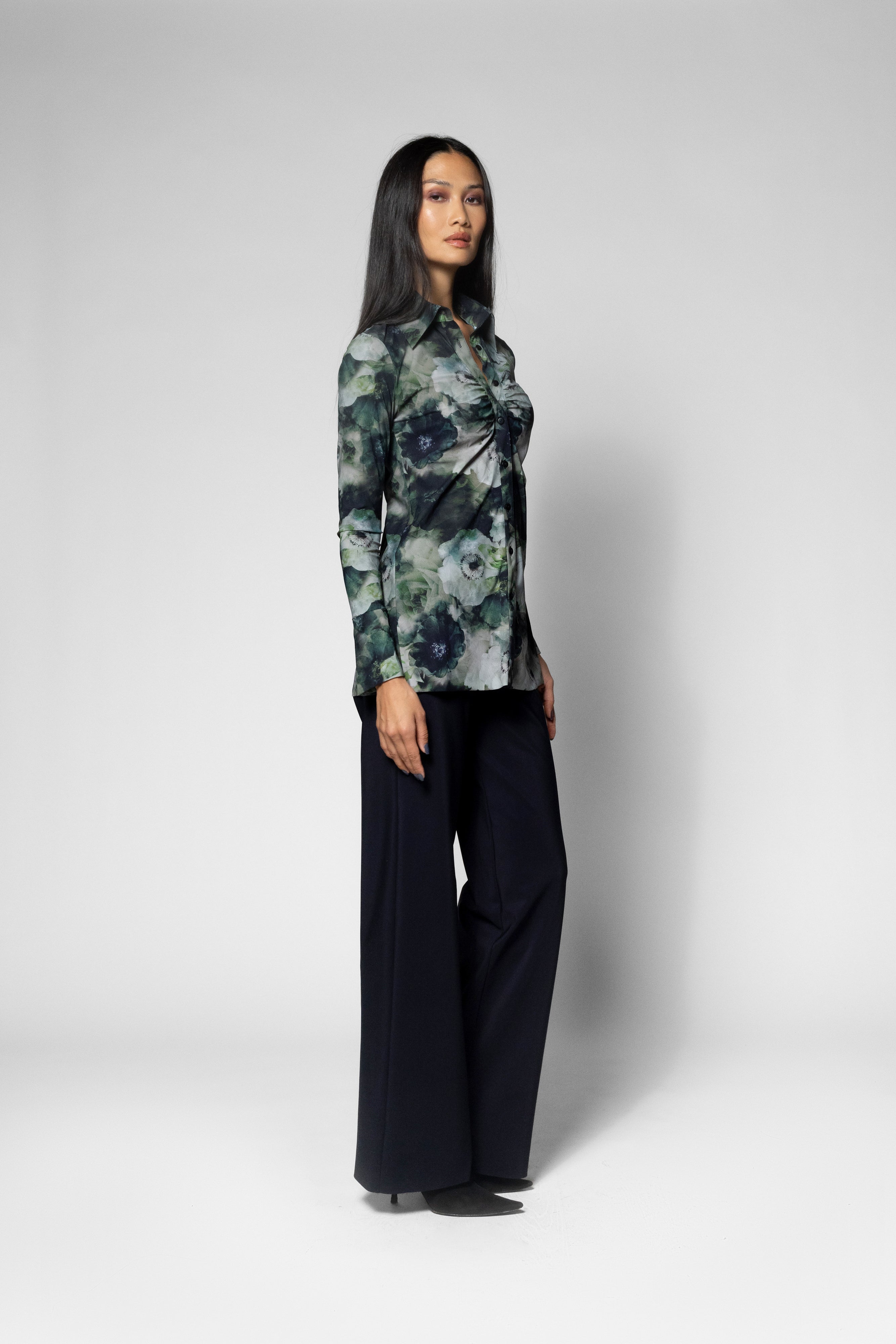 A woman modeling a long-sleeve button-up shirt with a green, blue, and gray floral pattern, paired with dark wide-leg trousers, against a light gray studio background.