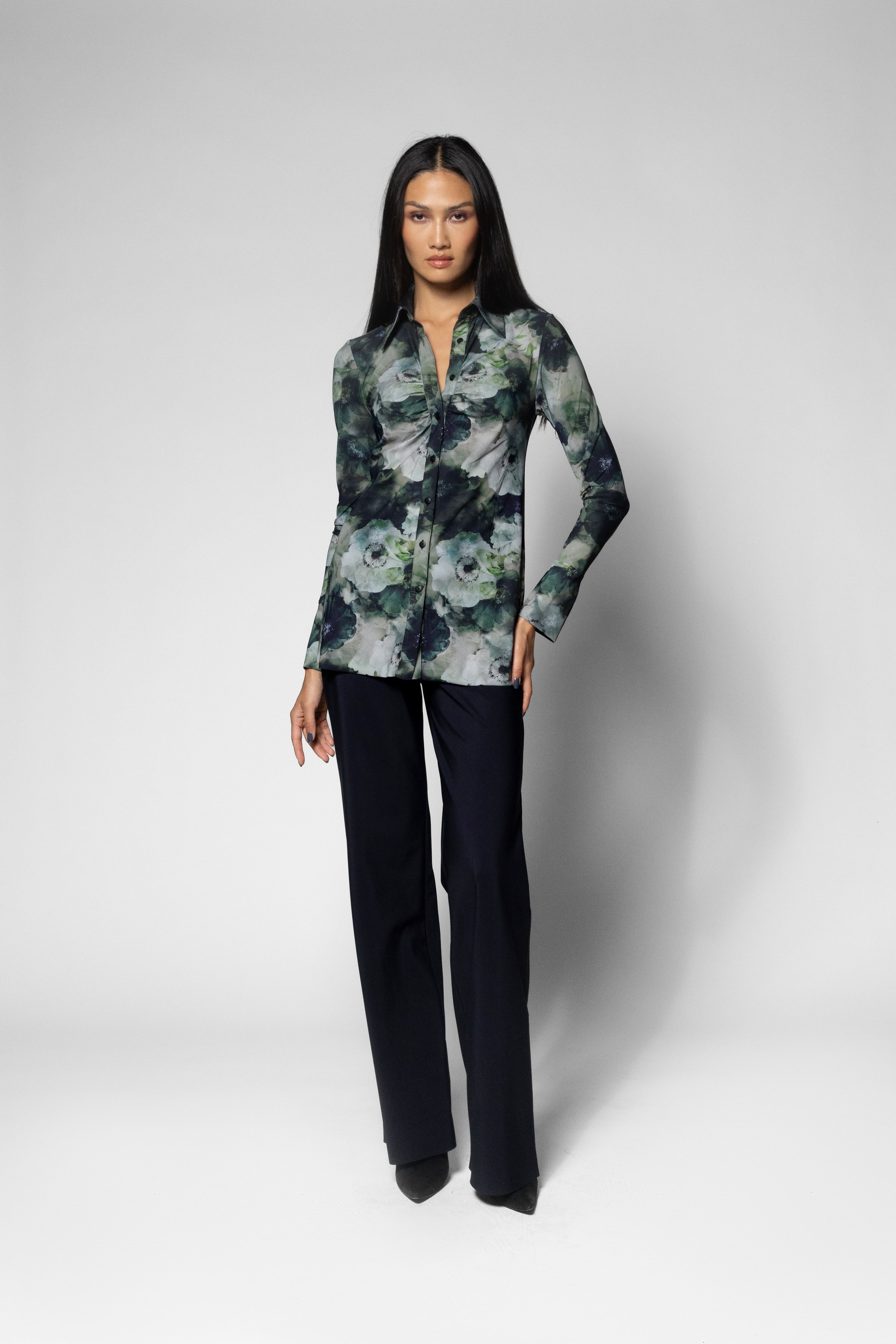 A woman modeling a long-sleeve button-up shirt with a green, gray, and black floral pattern, paired with dark trousers, against a light gray studio background.