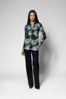 A woman modeling a long-sleeve button-up shirt with a green, gray, and black floral pattern, paired with dark trousers, against a light gray studio background.
