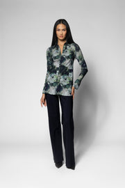 A woman modeling a long-sleeve button-up shirt with a green, gray, and black floral pattern, paired with dark trousers, against a light gray studio background.