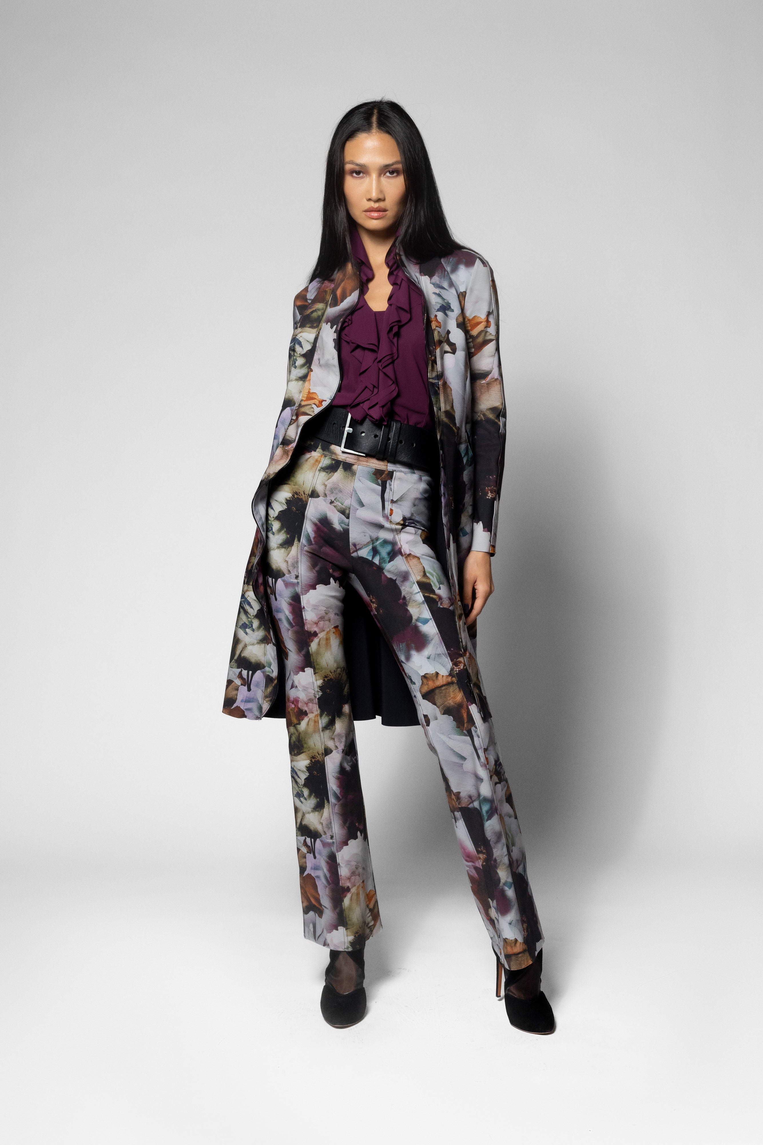 A model stands against a light gray studio background wearing a matching floral print blazer and trousers set with a dark purple ruffled blouse and black shoes.