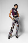 A model poses in a studio wearing a sleeveless plum top, a large black bow at the neckline, and high-waisted wide-leg trousers with a multicolor painterly floral print. She is also wearing a gray and black patterned scarf and black heels, set against a light gray background.