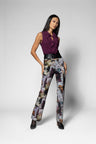 A model stands against a light gray studio background wearing a sleeveless dark purple ruffled top, a black belt, and high-waisted wide-leg trousers with a multicolor abstract floral print in purple, gray, beige, and brown. She is wearing black pointed-toe heels.
