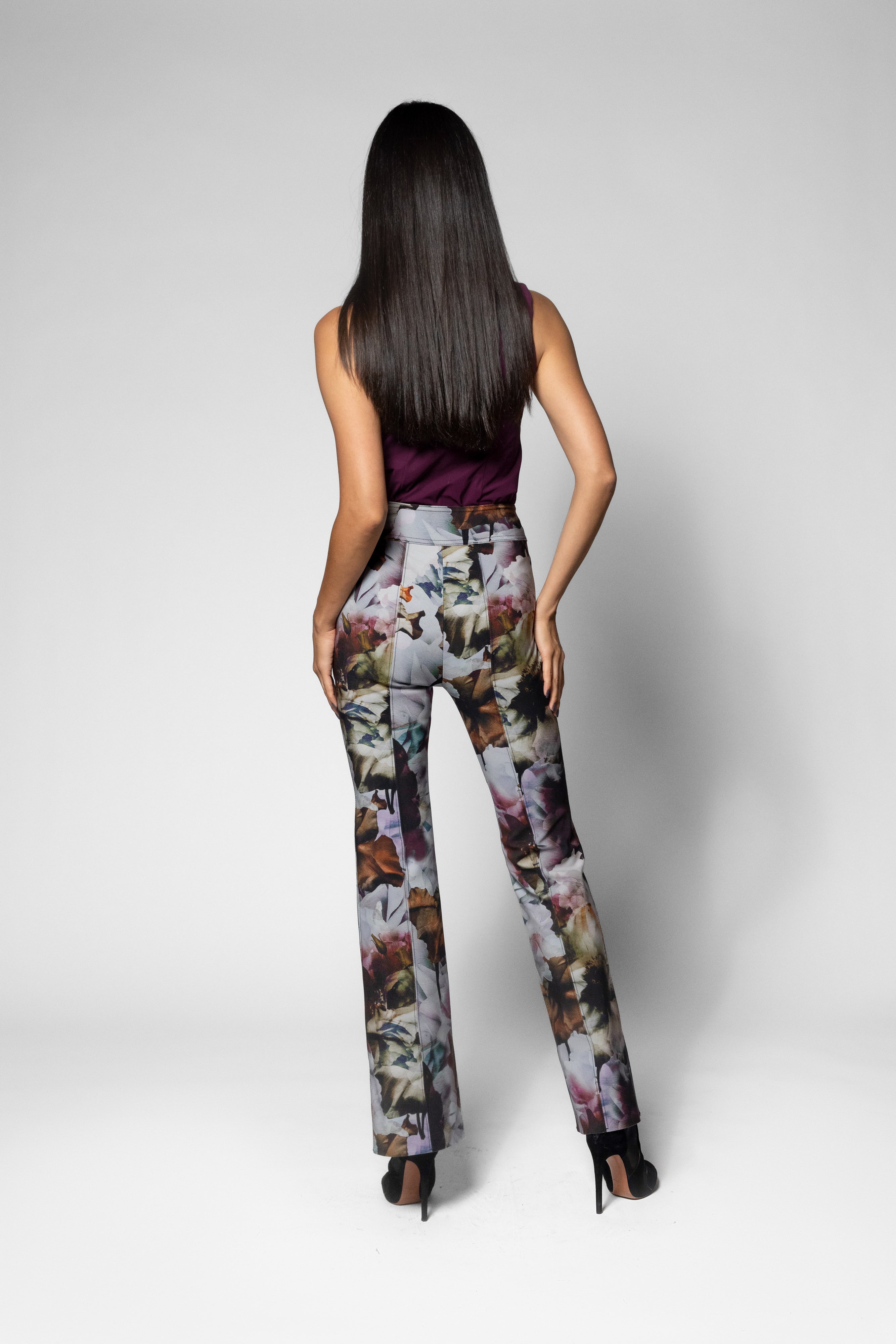 A woman shown from the back wearing high-waisted, wide-leg trousers with a multicolor floral and butterfly print, paired with a dark sleeveless top and black heels, against a light gray studio background.