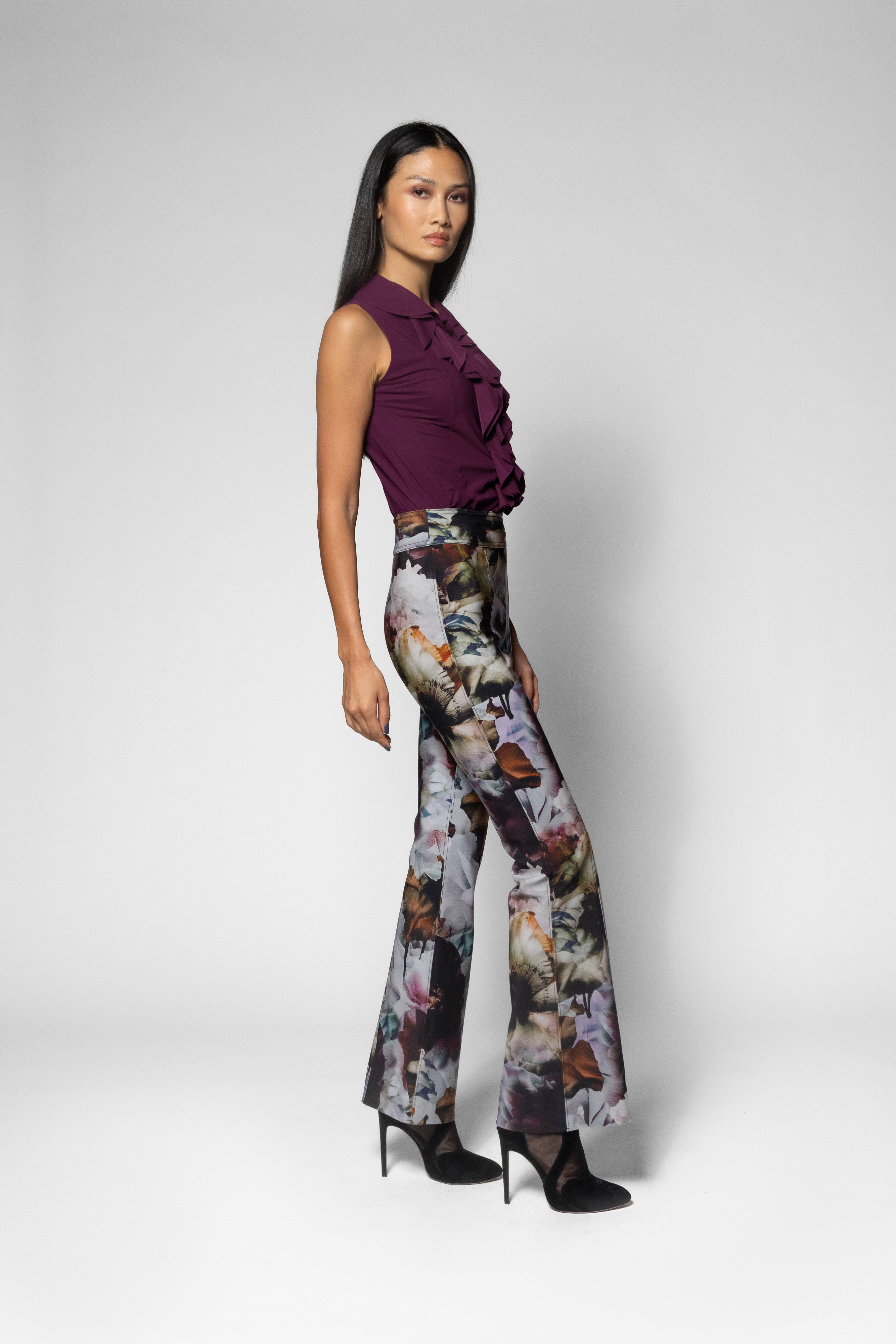 A model is shown from the side in a studio setting, wearing a sleeveless deep purple ruffled top and high-waisted, wide-leg trousers featuring a detailed butterfly and floral print in muted tones of gray, brown, and purple. She is wearing black high heels against a light gray background.