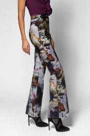 A model wearing high-waisted, wide-leg trousers with a multicolor abstract floral print, paired with black high heels and a dark purple top, shown from the waist down against a light gray studio background.