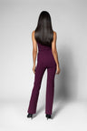 A woman with long dark hair is shown from the back wearing a sleeveless high-waisted pant in deep plum purple, paired with black heels, against a light gray studio background.