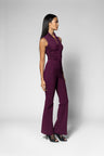 A woman modeling a sleeveless, high-waisted plum purple shirt with a wrap-style neckline and wide-leg pants, shown in profile against a light gray studio background.